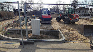 Baustelle mit Bauzaun, Bagger und Radlader auf sandigem Boden, im Hintergrund Häuser und Bäume