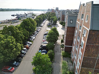 Blick von oben auf eine Straße mit parkenden Autos und Bäumen entlang eines Gewässers in einer Stadt.