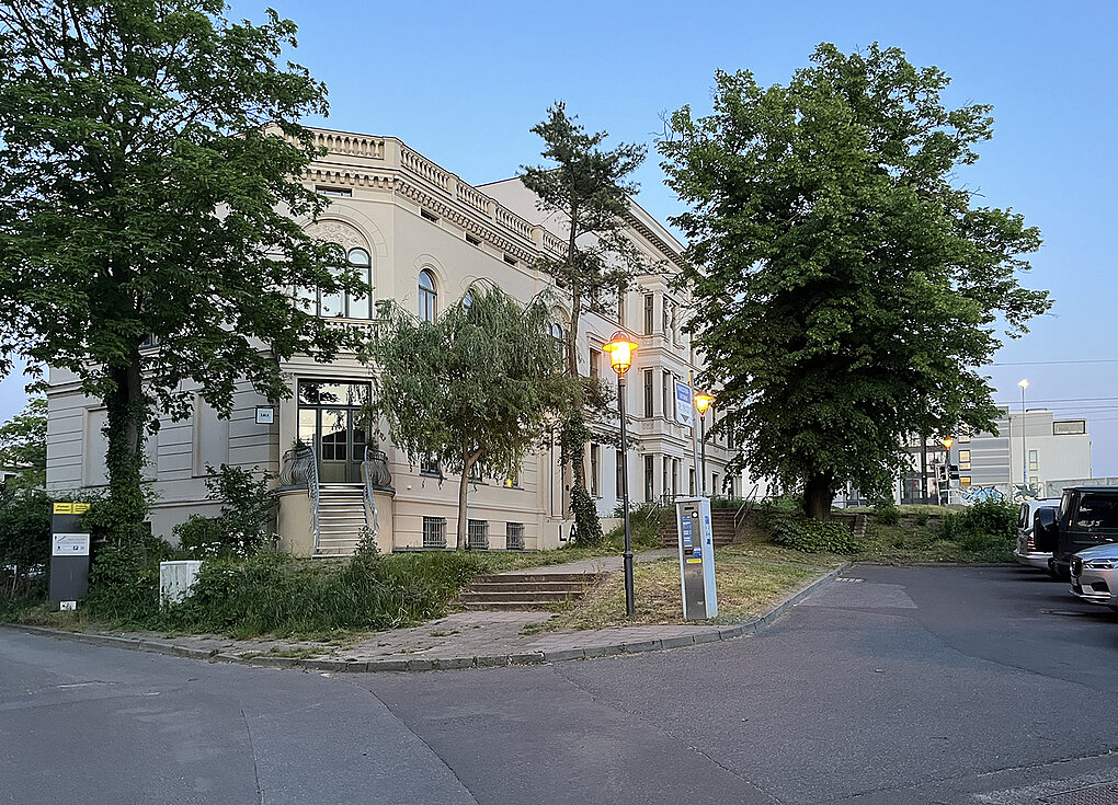 Eckhaus mit grüner Bepflanzung und Straßenlaterne, umgeben von Bäumen, bei Dämmerung.