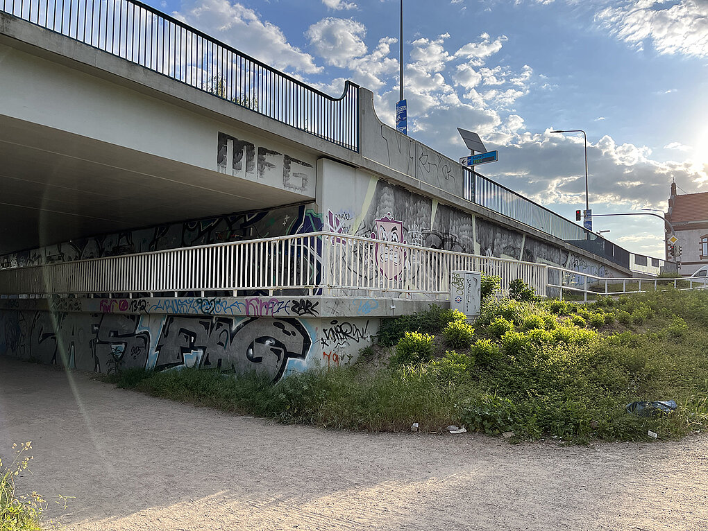 Unter einer Brücke sind Graffiti an der Wand sichtbar, umgeben von Grünpflanzen und Kies. Der Himmel ist blau mit einigen Wolken.