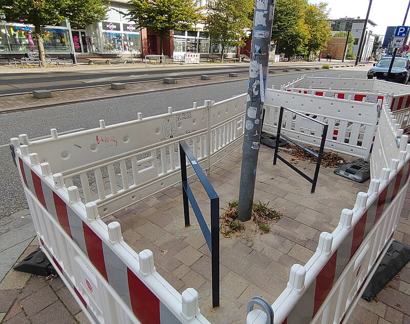 Zwei schwarze Fahrradbügel auf gepflastertem Boden umgeben von weißen Bauzäunen mit roten Streifen an einer Straßenbahnhaltestelle