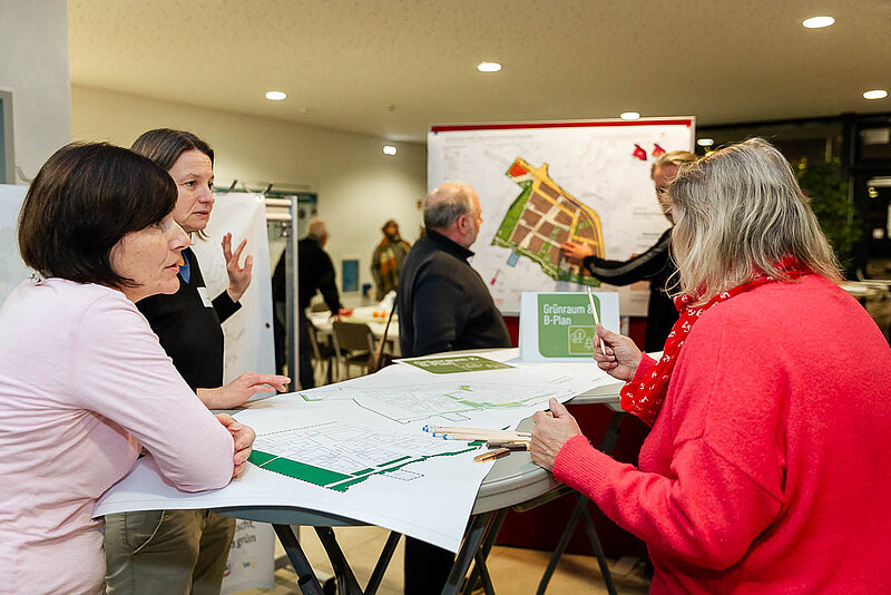 Vier Personen stehen an einem Tisch und diskutieren bei Plänen die auf dem Tisch liegen.