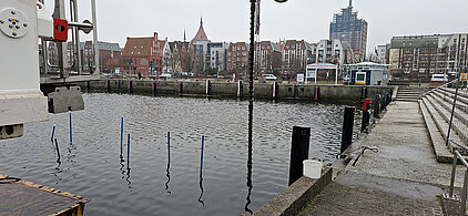 Hafenbecken mit Wasser, umgeben von Gebäuden und einem Kirchturm im Hintergrund, Uferpromenade mit Treppen und Pollern