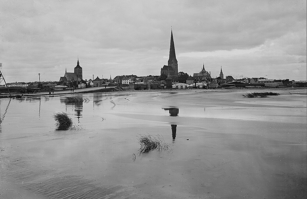Schwarz-weiß-Aufnahme von Rostock um 1930 mit mehreren Kirchtürmen, die sich in einem großen, ruhigen Gewässer spiegeln, unter bewölktem Himmel