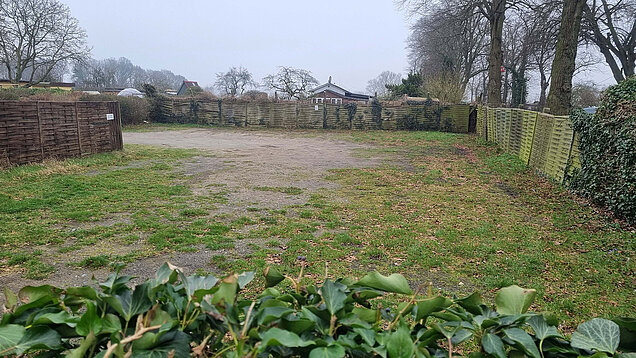 Leerer, unebener Garten mit Gras und Erde, umgeben von Holzzäunen und Bäumen im Hintergrund