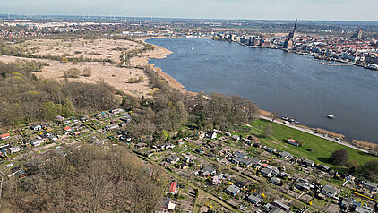Luftaufnahme der Unterwarnow mit der Stadt Rostock am Ufer und einem großen freien Fläche neben einem Gartenkolonie