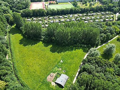 Luftaufnahme eines Parks mit großen Wiesenflächen, einem Gehweg und einem kleinen Gebäude, umgeben von Bäumen und einer Gartenanlage im Hintergrund