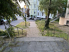 Blick auf einen Gehweg, der von Bäumen gesäumt ist, mit einem schmalen Pfad, der zu einer Straße führt. Im Hintergrund sind Wohngebäude und parkende Autos sichtbar.