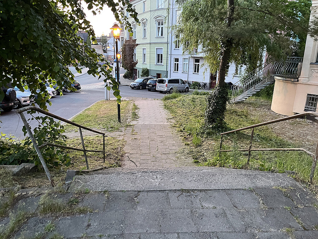 Blick auf einen Gehweg, der von Bäumen gesäumt ist, mit einem schmalen Pfad, der zu einer Straße führt. Im Hintergrund sind Wohngebäude und parkende Autos sichtbar.