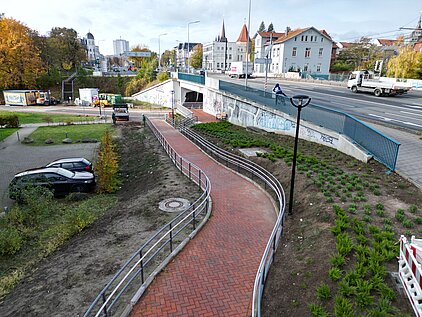 Luftaufnahme eines gepflasterten, geschwungenen Fußwegs mit Geländer neben einer Straße und einem Parkplatz in einer Stadt mit herbstlichen Bäumen