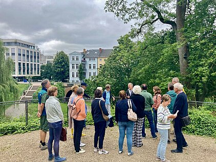 Gruppe von Menschen, die in einem Park stehen und sich umschauen, im Hintergrund moderne Gebäude und Bäume.