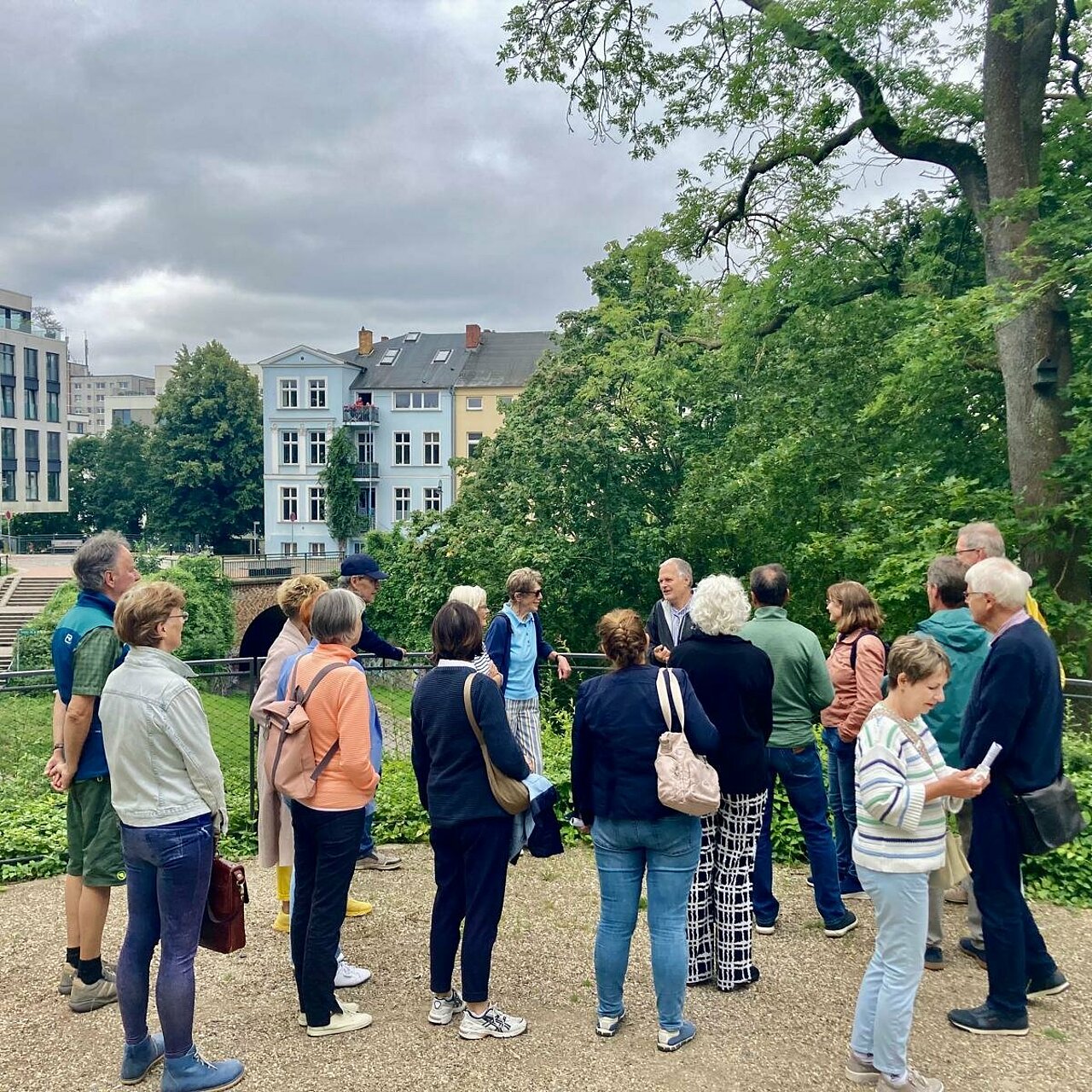 Gruppe von Menschen, die in einem Park stehen und sich umschauen, im Hintergrund moderne Gebäude und Bäume.