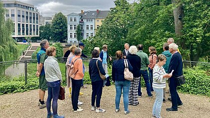 Gruppe von Menschen, die in einem Park stehen und sich umschauen, im Hintergrund moderne Gebäude und Bäume.