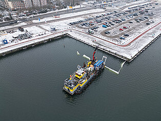 Schiff mit Kran auf Wasserfläche vor schneebedecktem Hafen mit Parkplätzen und Gebäuden im Hintergrund