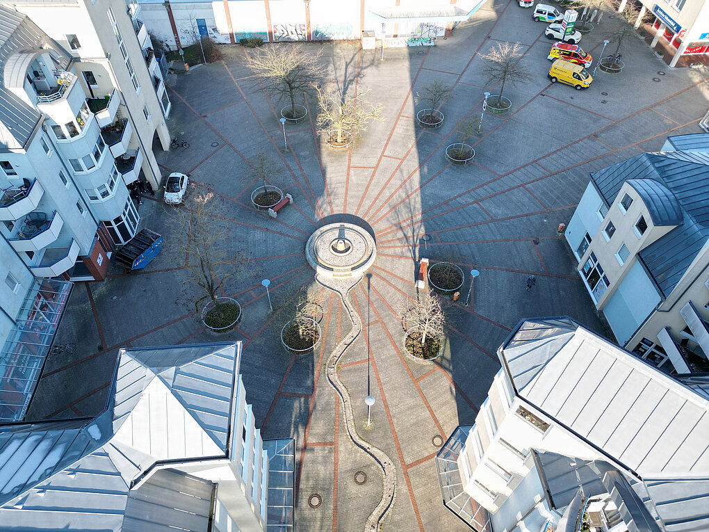Luftaufnahme eines Platzes mit einem runden Brunnen in der Mitte, umgeben von Gebäuden