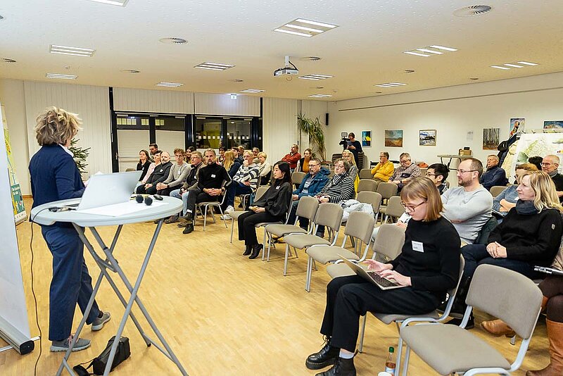 Ein Raum voller sitzender Menschen. Im Vordergrund eine Frau die spricht.