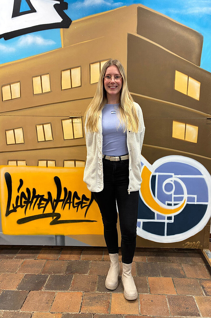 Frau mit langen blonden Haaren, hellblauem Oberteil, weißer Strickjacke, schwarzer Hose und hellen Stiefeln steht vor einer bunten Wand mit Graffiti und stilisierten Gebäuden