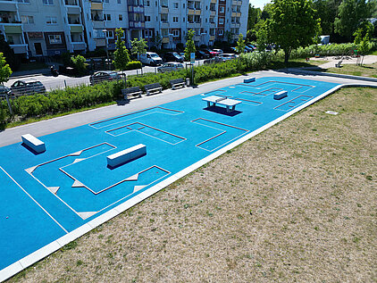 Minigolfbahn mit blauer Spielfläche und verschiedenen Hindernissen im Freien, umgeben von Gras und Wohngebäuden im Hintergrund