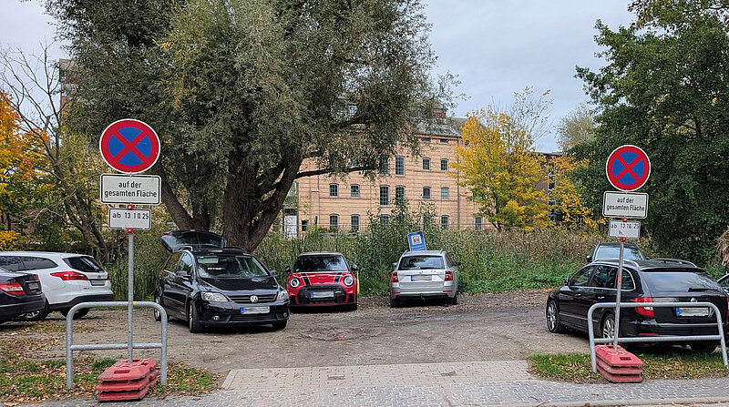 Parkplatz mit mehreren geparkten Autos, zwei Verkehrsschilder mit rotem Kreuz auf blauem Grund und Zusatzschildern 'auf der gesamten Fläche' und 'ab 13.10.25' vor Bäumen und einem mehrstöckigen Gebäude