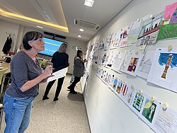 Drei Personen betrachten eine Wand mit zahlreichen bunten Zeichnungen und Illustrationen in einem hellen Raum.