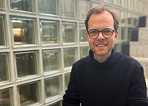 Mann mit Brille und dunklem Pullover, lächelt vor einer Wand aus Glasbausteinen.