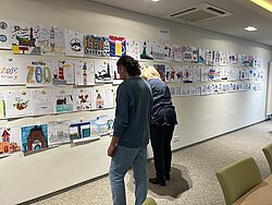Zwei Personen betrachten an einer Wand zahlreiche bunte Zeichnungen und Bilder, die in Reihen angeordnet sind.