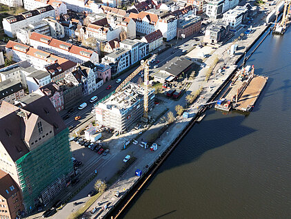 Luftaufnahme eines Stadthafens mit Baustelle, Kran, parkenden Autos und angrenzenden Wohngebäuden am Flussufer