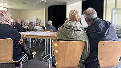Menschen sitzen auf Stühlen in einem Raum und schauen auf eine Präsentationsleinwand