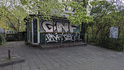 Kleines Gebäude mit Graffiti an den Wänden, umgeben von Bäumen und gepflastertem Platz
