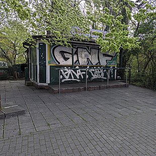 Kleines Gebäude mit Graffiti an den Wänden, umgeben von Bäumen und gepflastertem Platz
