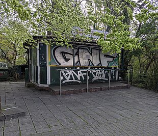 Kleines Gebäude mit Graffiti an den Wänden, umgeben von Bäumen und gepflastertem Platz