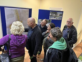 Gruppe von Menschen schaut auf eine Stellwand an der Baupläne hängen und diskutieren.