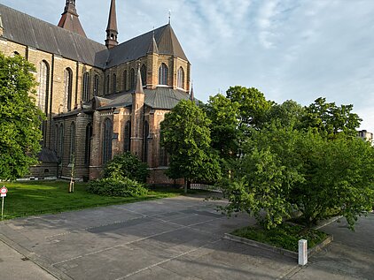 Gotische Kirche mit spitzem Turm neben einem gepflasterten Weg und grünen Bäumen bei klarem Himmel.