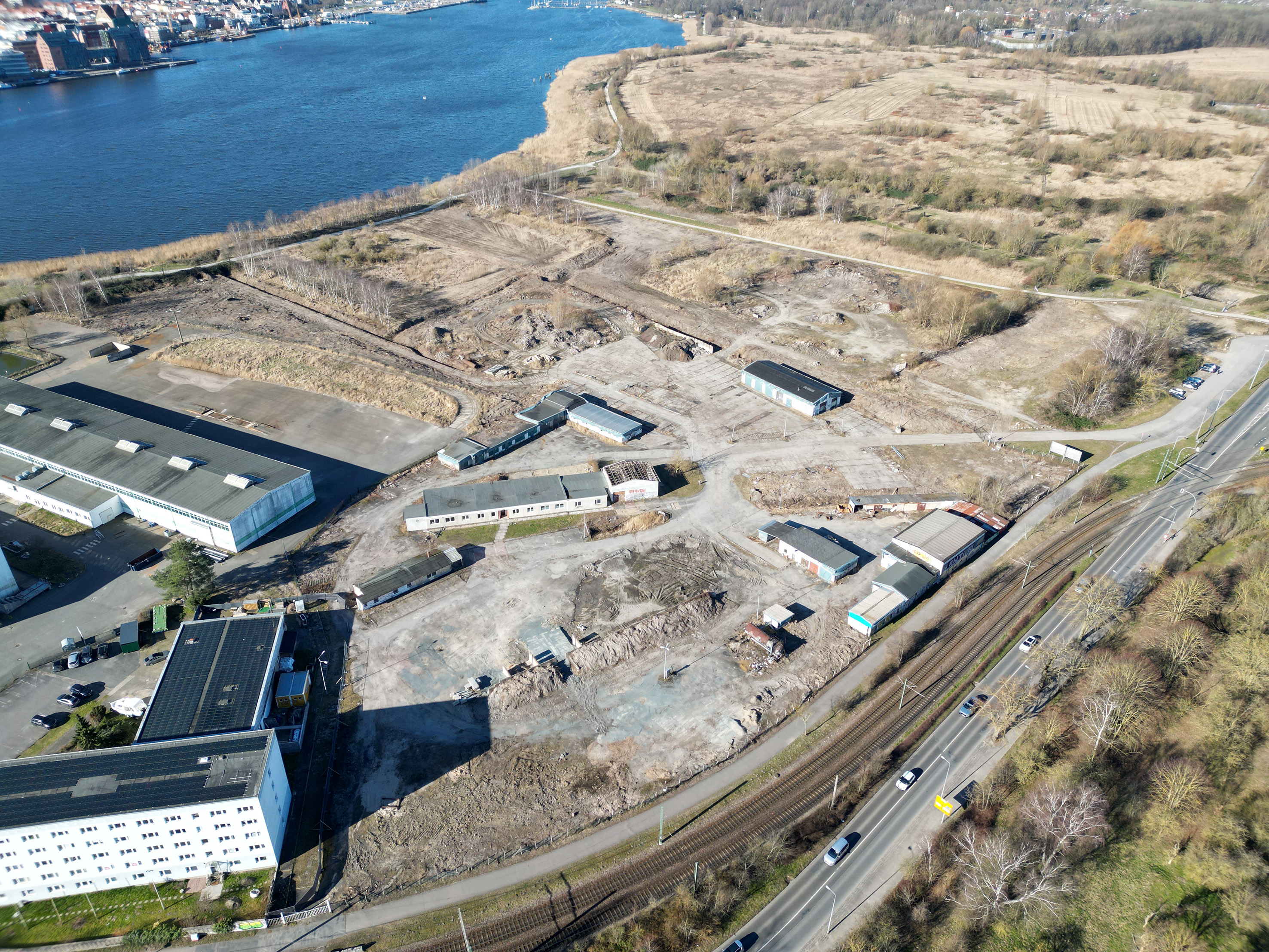 Luftaufnahme des Geländes des künftigen WarnowQuartiers mit Blick auf die ungenutzten Gebäude des ehemaligen Bauhofes, umgeben von Wasser und Straßen. Im Hintergrund sind Gebäude und eine Stadt sichtbar.