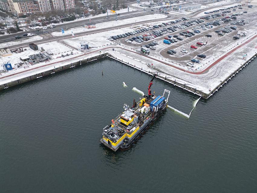 Schiff mit Kran auf Wasserfläche vor schneebedecktem Hafen mit Parkplätzen und Gebäuden im Hintergrund