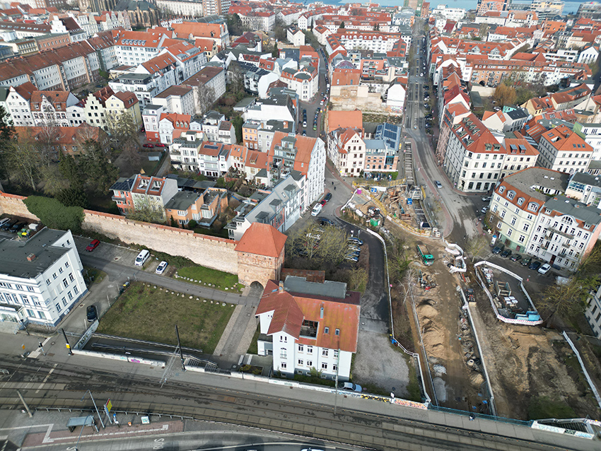 Luftaufnahme von Rostock mit roten Dächern, Baustelle im Vordergrund und Straßenbahnschienen im unteren Bildbereich.