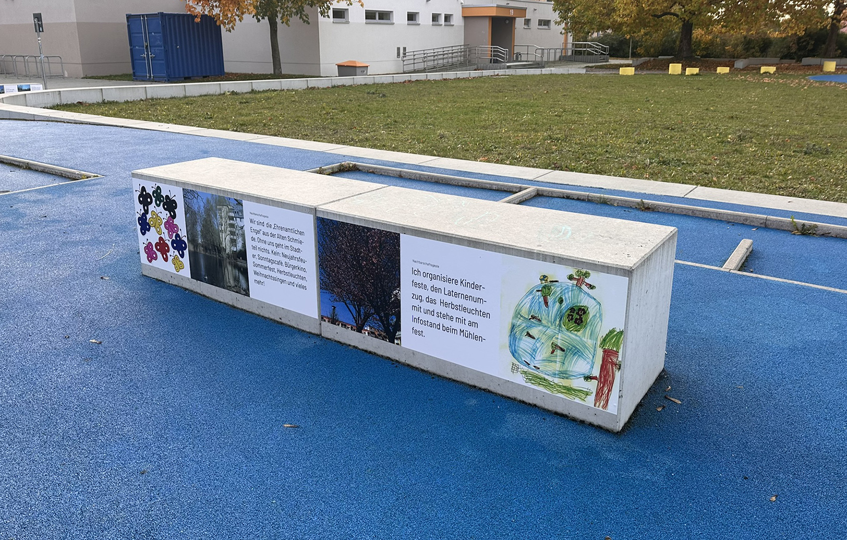 Betonbank mit vier aufgeklebten Bildern und Texten auf der Seite auf blauem Boden vor einer Wiese und Gebäuden